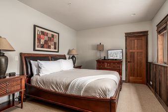 A king bedroom at Latigo Lodge featuring a wooden bed frame, white linens, two nightstands with lamps, a TV on a dresser, a wooden door, and a rustic window.