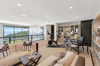 A well-lit room features a comfortable lounge area, a dedicated workspace with a desk and globe, and a fitness corner with a stationary bike, all set against a backdrop of large windows offering scenic mountain and water views.