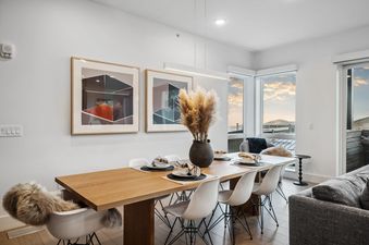 A modern dining room in Apex 208 features a wooden table set for six, surrounded by white chairs, with abstract art on the wall and large windows offering scenic outdoor views.