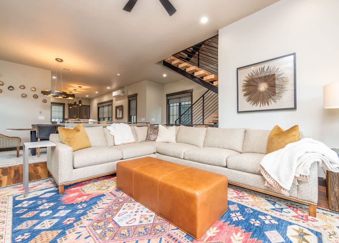 An expansive open-concept living area at Sky High Colorado with a large L-shaped sectional sofa, a brown leather ottoman, and a colorful patterned rug. In the background, a dining area and a modern staircase are visible.