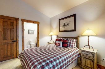 A bedroom with a plaid comforter and a wooden bed frame. There is a nightstand with a lamp next to the bed, and a door to a bathroom on the left.