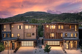 The Yampa Vista Grande features a sleek, modern architectural design with large windows and an inviting driveway, set against a stunning mountain backdrop with a vibrant sunset sky.