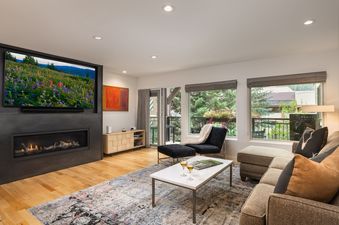 The living room of Montaneros 214 features a modern fireplace with a television above, a comfortable sectional sofa, and large windows with views of greenery.