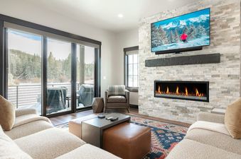 A modern living room with a large sectional sofa, a stone fireplace, a wall-mounted TV displaying a skiing scene, and a balcony with a snowy mountain view.