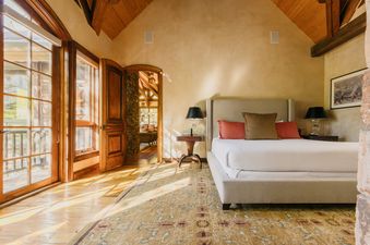A sunlit king bedroom at Four Bears Hideaway with large glass doors opening to a balcony, a plush bed with colorful pillows, and rustic wooden accents.