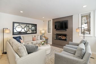 The comfortable and stylish living room of Tamarack 04, featuring a sectional sofa, armchairs, a fireplace, and a television, bathed in natural light from windows overlooking a snowy outdoor scene.