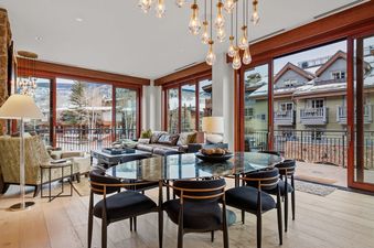A luxurious open-plan living and dining area in Solaris 2A South, featuring a stylish dining table, elegant chandelier, comfortable leather sofa, and expansive glass doors leading to a balcony with mountain views.