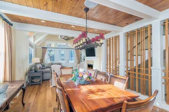 A spacious dining area featuring a large wooden table, an ornate chandelier, and an adjacent living room with a fireplace and outdoor views.