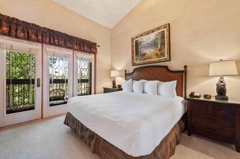 A spacious bedroom in The Charter C2205 features a king-size bed with a brown leather headboard, white linens, and a plaid bed skirt, with an elegant landscape painting above it and a nightstand with a bear-shaped lamp.