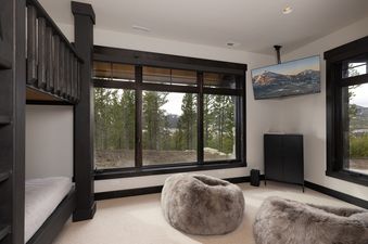A cozy bedroom at Summit View Chalet Big Sky with a bunk bed, two plush bean bags, and a flat-screen TV, featuring large windows that showcase a stunning view of a pine forest and distant mountains.