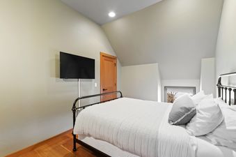 A cozy bedroom features a black wrought iron bed with white and grey striped bedding, a wall-mounted flat-screen TV, and warm hardwood floors.