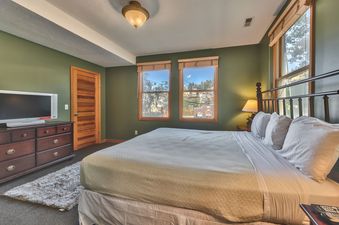 A serene bedroom with a large bed, olive green walls, and two windows offering a view of the neighborhood.