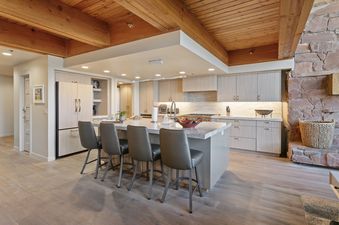 A modern kitchen at Stag Lodge 10 with light wood cabinetry, a large marble island with four gray bar stools, and a contemporary linear chandelier overhead.