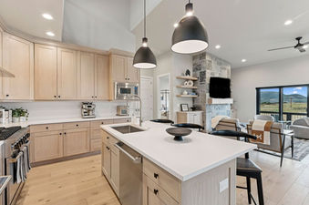 A modern kitchen with light wood cabinetry, white countertops, and stainless steel appliances, seamlessly connecting to a living area with a stone fireplace and mountain views at Deer Valley Vista.