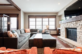 A cozy living room at Silver Star 1304 featuring an L-shaped sectional sofa with throw pillows, a large ottoman acting as a coffee table, and a stone fireplace with a mounted flat-screen TV.