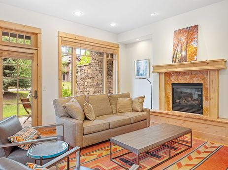 A cozy living room featuring a fireplace, a comfortable sofa, armchairs, and a vibrant geometric rug, with a large window and glass-paneled door offering views of the outdoors.
