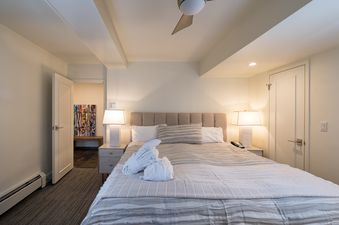 A cozy bedroom with a large bed, two nightstands with lamps, and an open doorway leading to another room with colorful art.