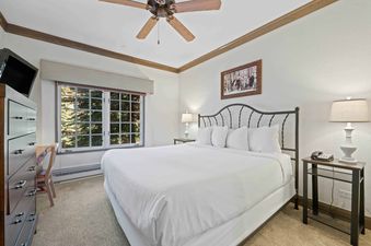 A cozy bedroom in The Charter E0205 featuring a large bed with white linens, a metal headboard, bedside tables with lamps, a desk with a chair, a television, and a window overlooking a lush outdoor scene.