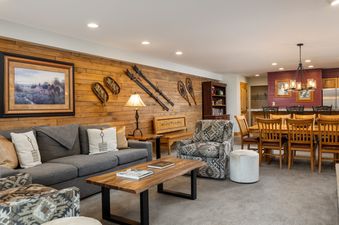 An inviting open-plan living and dining area with rustic wood paneling, comfortable seating, and a large dining table under a stylish chandelier.