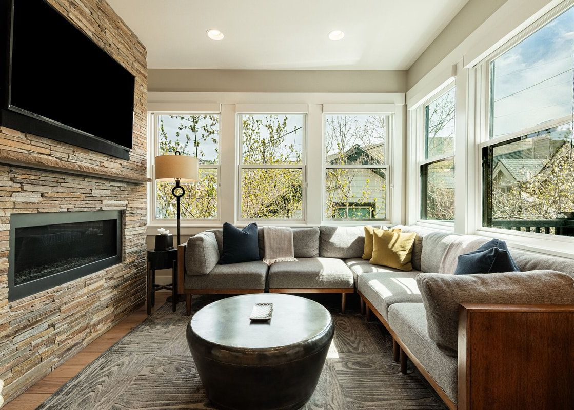 A cozy living room with a grey sectional sofa, a stacked-stone fireplace, and large windows looking outside.