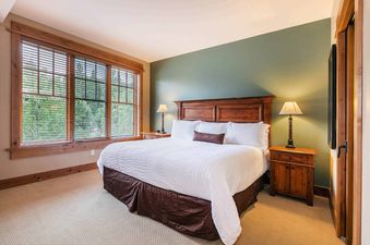 A bedroom with a large window, a king size bed with white sheets and a brown bed skirt, and two wooden bedside tables with lamps.