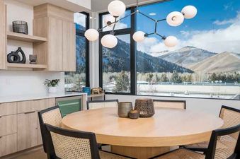 A dining room with a large window showcasing a mountain view, featuring a round wooden table with four chairs, a modern light fixture, and a kitchen counter with cabinets.