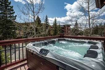 A private outdoor hot tub on the wooden deck of Trademark Views, surrounded by evergreen and aspen trees under a partly cloudy sky.