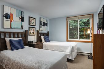 A peaceful twin bedroom in Terracehouse 63 features two beds with white bedding, navy blue pillows, a dark wood nightstand, abstract art, and a window with a green exterior view.