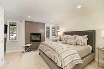 A serene bedroom in Tamarack 04 featuring a king-size bed with striped bedding, a modern fireplace, and windows revealing a snowy exterior view.