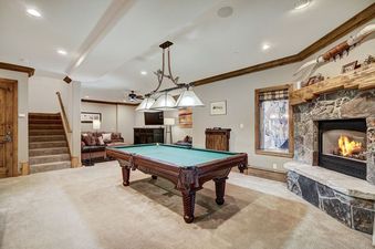 The game room at Arrowhead Village Condominium Platinum 4 Bedroom and Den features a full-sized billiard table under decorative lighting, with a cozy seating area and a stone fireplace nearby.