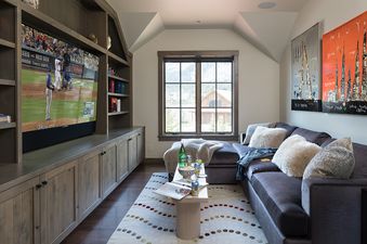 A cozy entertainment area with a large TV showing a baseball game, a comfortable sectional sofa with pillows, and a window overlooking an outdoor view.