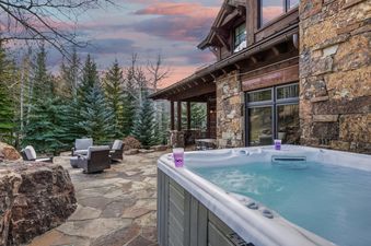 The hot tub at the High Road property is shown on a stone patio with outdoor seating, surrounded by trees under a sunset sky, next to the stone and timber house.