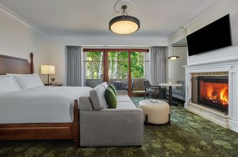 The Montage Deer Valley King Room Resort View features a luxurious king-sized bed, a comfortable sofa, and a cozy fireplace, all set against a backdrop of natural beauty visible through large windows leading to a private balcony.