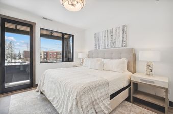 A king-sized bed with white bedding, tufted headboard, and birch tree artwork, flanked by a nightstand with a lamp and a balcony door and window with winter views.