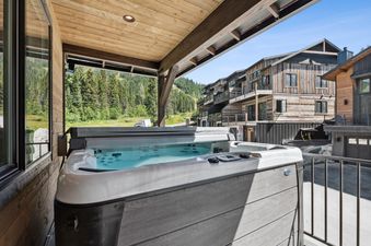 The hot tub at Sweet Peaks, nestled under a wooden patio with a view of a lush forest, mountains, and elegant wooden buildings.