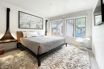 A bedroom with a plush gray carpet and a plush gray duvet on the bed. The room has a large window overlooking the forest.