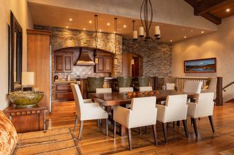The dining area at Lynx Lookout at See Forever features a long wooden table with upholstered chairs, set against a backdrop of a rustic stone kitchen with wooden cabinets.