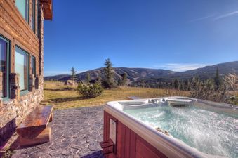 A hot tub with bubbling water sits on a stone patio outside a rustic lodge, overlooking a mountain landscape under a clear blue sky.