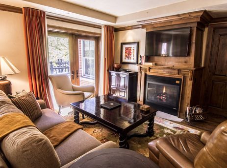 A cozy living room at the Hyatt 3 Bedroom features a comfortable sofa, a large coffee table, and a fireplace with a flat-screen TV above it, all illuminated by a table lamp.