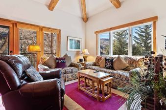 The living area at The Charter E2205 features plush floral sofas, a comfortable leather armchair, and a wooden coffee table on a magenta rug, all illuminated by large windows with scenic mountain views.