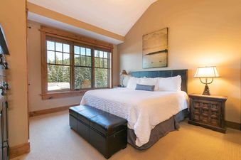 A bedroom with a white bed, a black leather bench at the foot of the bed, and a wooden nightstand with a lamp on it. There is a window with a view of a snowy forest.