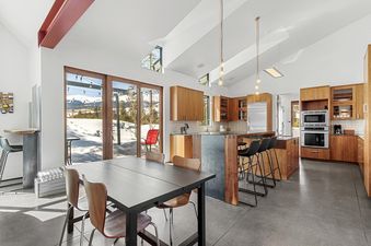 The open-concept kitchen and dining area of the Modern Alpine Escape, featuring wooden cabinetry, stainless steel appliances, a dining table, and large glass doors revealing a snow-covered mountain view.