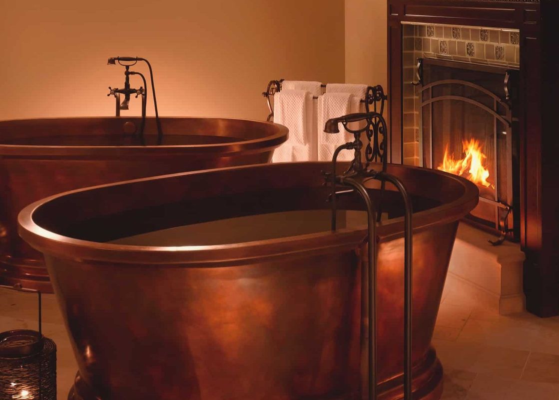 Two large, opulent copper bathtubs filled with water stand side by side in a dimly lit, warm-toned bathroom, with a glowing fireplace in the background.