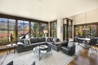 A spacious living and dining area in On Trails Edge, featuring a gray sectional sofa, a round dining table, and large windows with mountain views.