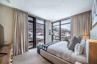 The bedroom in One Snowmass Residence Club 407 features a large bed with a plaid throw and multiple pillows, facing floor-to-ceiling windows with panoramic views of snow-covered mountains.