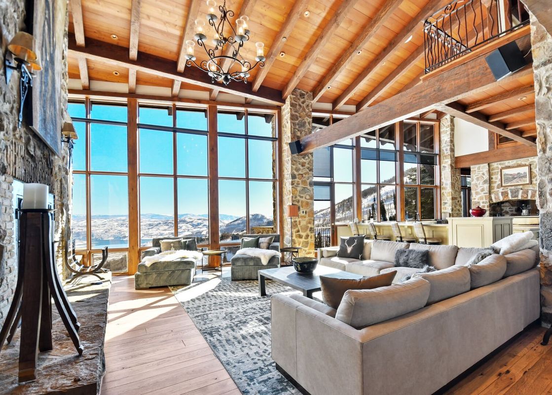 The Snow Pine Lodge at Deer Valley great room showcases a vaulted wood-paneled ceiling, a large chandelier, and floor-to-ceiling windows with panoramic views of snow-covered mountains and a lake, complemented by plush sofas and a stone fireplace.