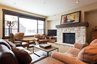 A spacious and inviting living room at Silver Star 4202 features a stone fireplace, comfortable seating, a flat-screen TV, and large windows with a view of a snowy landscape.