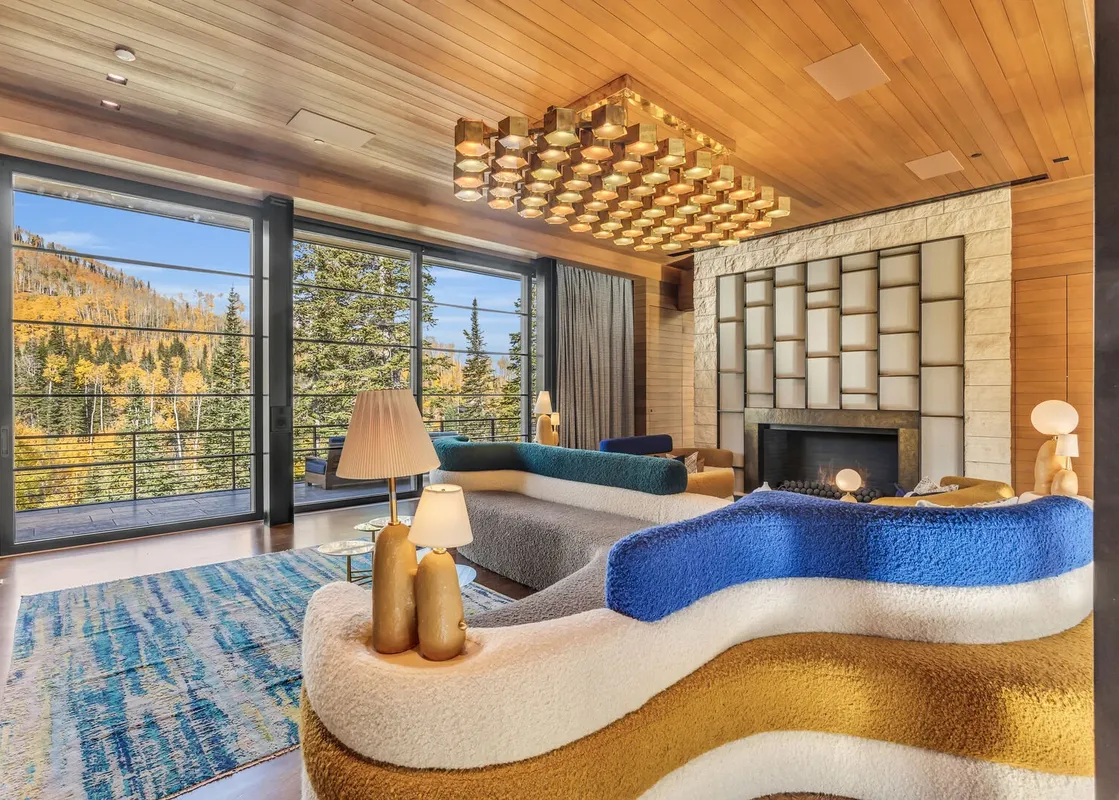 The living area of Monitors Rest features a vibrant, multi-layered sofa, a distinctive gold-toned chandelier, a modern fireplace, and expansive windows with breathtaking views of an autumnal forest.