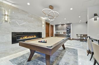 A luxurious games room with a billiards table, a modern fireplace set into a marble wall, a wet bar, and contemporary lighting.