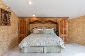 A cozy bedroom with a large bed featuring a wooden headboard, light blue and green patterned bedding, and a horse painting on the mustard yellow floral wallpaper.